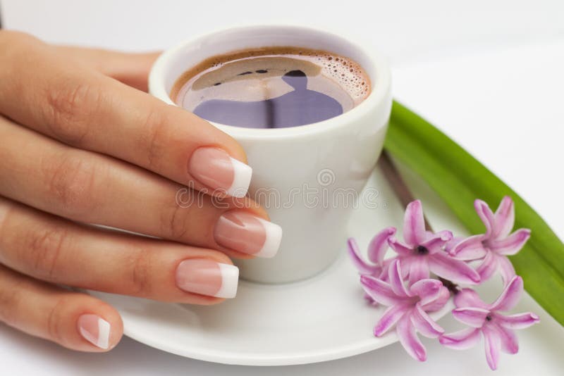 Όμορφος το χέρι με τα γαλλικά καρφιά και το φλιτζάνι του καφέ και τα λουλούδια στο πιατάκι