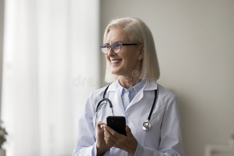 Mature female doctor standing at workplace with smartphone, accessing electronic health records, communicate with colleague or patient via call, texts, apps, setting reminder for appointment or task. Mature female doctor standing at workplace with smartphone, accessing electronic health records, communicate with colleague or patient via call, texts, apps, setting reminder for appointment or task