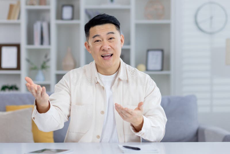 Cheerful Asian man engaging in a casual conversation at home, looking at the camera and talking with expressive hand gestures in a modern living room setting. Cheerful Asian man engaging in a casual conversation at home, looking at the camera and talking with expressive hand gestures in a modern living room setting.