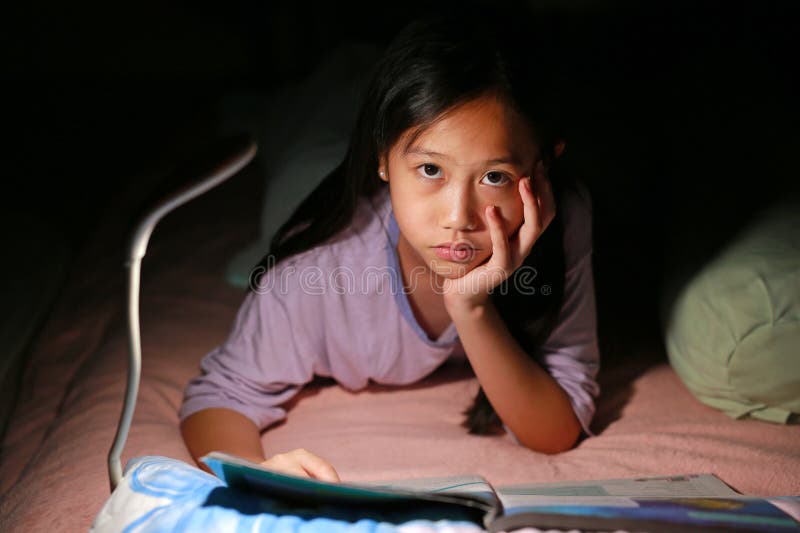 Азиатская девочка читает книгу с лампой, лежа ночью на кровати. Приблизительный снимок стоковое изображение