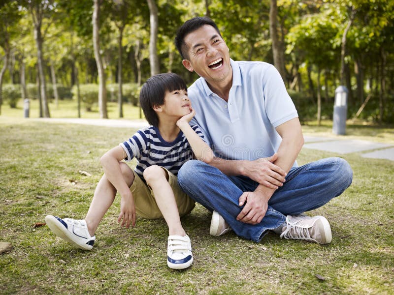 Asian father and elementary-age son sitting on grass outdoors having an interesting conversation. Asian father and elementary-age son sitting on grass outdoors having an interesting conversation.