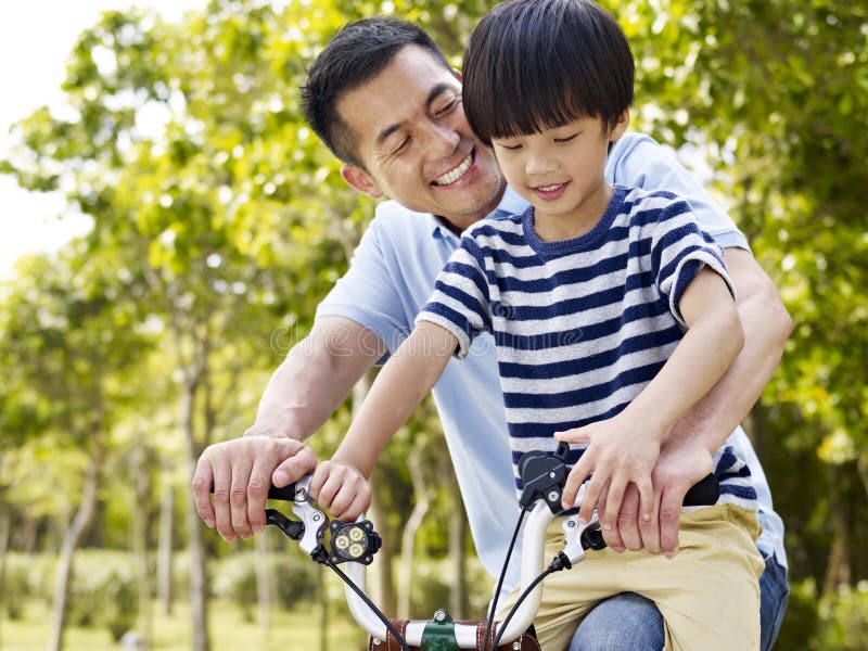 Asian father and elementary-age son enjoying riding a bike outdoors in a park. Asian father and elementary-age son enjoying riding a bike outdoors in a park.