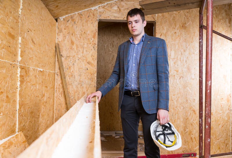 Portrait of Young Male Architect Holding White Hard Hat and Wearing Suit Jacket Standing Inside Unfinished Home with Exposed Particle Plywood Board. Portrait of Young Male Architect Holding White Hard Hat and Wearing Suit Jacket Standing Inside Unfinished Home with Exposed Particle Plywood Board