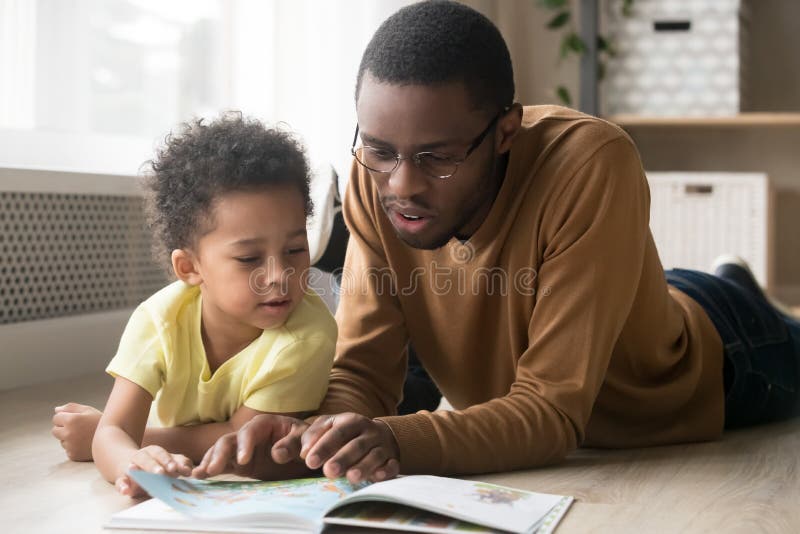 African American father in glasses and toddler son reading book together, lying on warm floor at home, black dad close up reading aloud fairy tale to adorable child, education, family weekend. African American father in glasses and toddler son reading book together, lying on warm floor at home, black dad close up reading aloud fairy tale to adorable child, education, family weekend