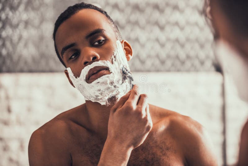 Young Afro American Man Shaving in Bathroom at Morning. Standing Man with Bare Torso in Bathroom. Personal Morning Routine. Fresh at Morning. Guy near Mirror. Hygiene at Morning Concept. Young Afro American Man Shaving in Bathroom at Morning. Standing Man with Bare Torso in Bathroom. Personal Morning Routine. Fresh at Morning. Guy near Mirror. Hygiene at Morning Concept.