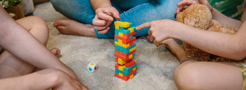 Banner of unrecognizable family having fun together playing with wooden stacking piece game over carpet at home. Little girl pointing with her finger to piece of game while holding a stuffed teddy dog. Banner of unrecognizable family having fun together playing with wooden stacking piece game over carpet at home. Little girl pointing with her finger to piece of game while holding a stuffed teddy dog