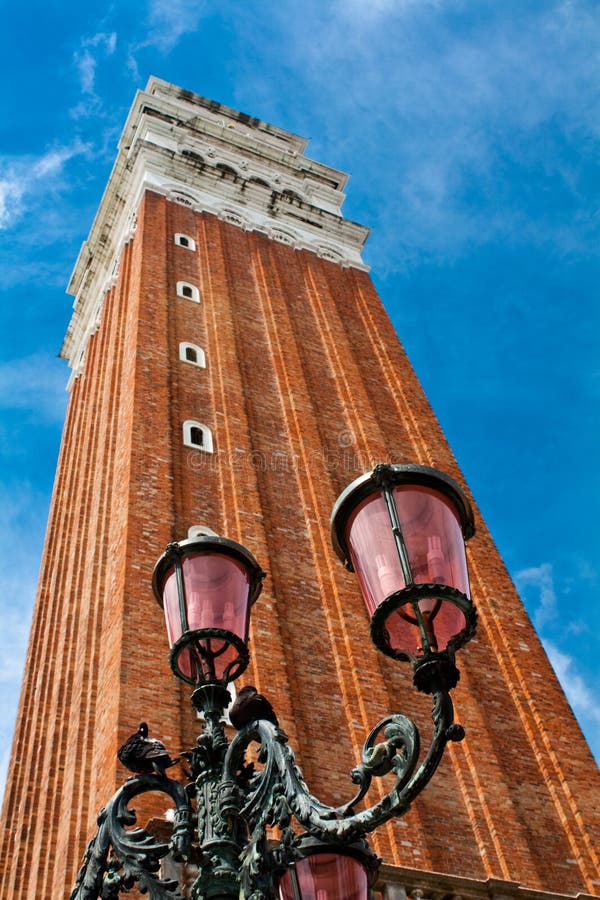 The bell tower from San Marco Square (St Mark's Campanile) in Venice, Italy. The tower is 98.6 meters (323 ft) tall, and stands alone in a corner of St Mark's Square. A street lamp with 2 pigeons is in the foreground. The bell tower from San Marco Square (St Mark's Campanile) in Venice, Italy. The tower is 98.6 meters (323 ft) tall, and stands alone in a corner of St Mark's Square. A street lamp with 2 pigeons is in the foreground.