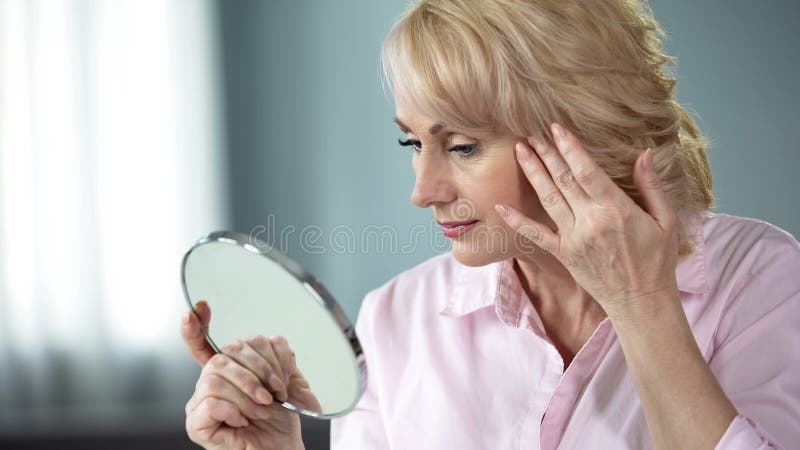 Blond middle-aged lady looking in mirror at her eye wrinkles, beauty injections, stock photo. Blond middle-aged lady looking in mirror at her eye wrinkles, beauty injections, stock photo