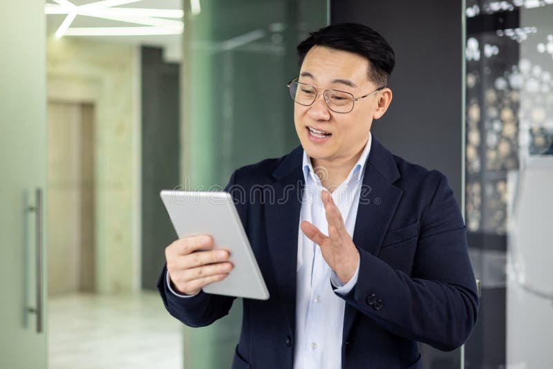 Confident Asian businessman communicating via a digital tablet in a modern office environment, gesturing with his hand, dressed in a smart business suit. Confident Asian businessman communicating via a digital tablet in a modern office environment, gesturing with his hand, dressed in a smart business suit.