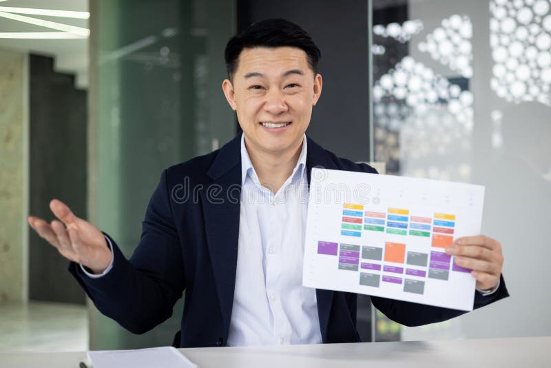 Confident Asian businessman discussing a colorful strategy chart while smiling and talking to the camera, conveying a sense of leadership and professionalism in a modern office environment. Confident Asian businessman discussing a colorful strategy chart while smiling and talking to the camera, conveying a sense of leadership and professionalism in a modern office environment.