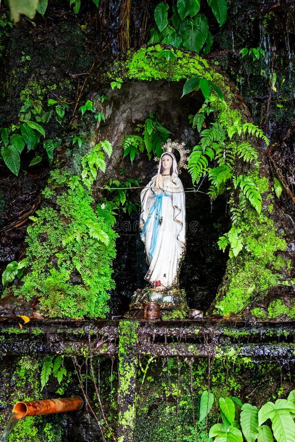 The virgin Mary statue surrounded by lush tropical vegetation such as sterns with a with fresh water pipe on a cliff falling into Pandin lake,  one of the seven lakes of San Pablo, Laguna Province, Luzon island, Philippines. The virgin Mary statue surrounded by lush tropical vegetation such as sterns with a with fresh water pipe on a cliff falling into Pandin lake,  one of the seven lakes of San Pablo, Laguna Province, Luzon island, Philippines