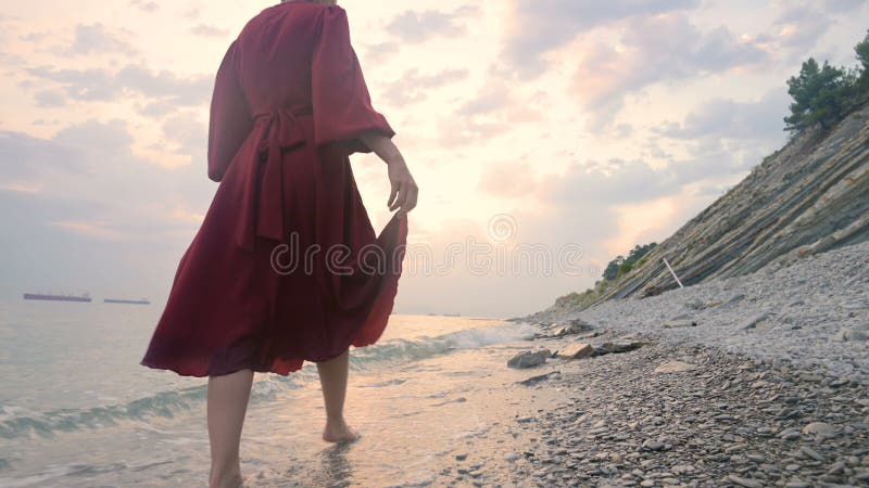 Близость руки молодой девушки с красным платьем из-за спины, идущей по скалистому пляжу на морском побережье на закате A