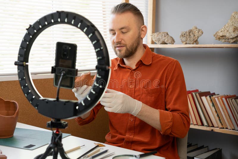 Archaeologist blogger broadcasts live with a ring lamp in his office working with ancient artifacts of prehistoric culture. Copy space. Archaeologist blogger broadcasts live with a ring lamp in his office working with ancient artifacts of prehistoric culture. Copy space