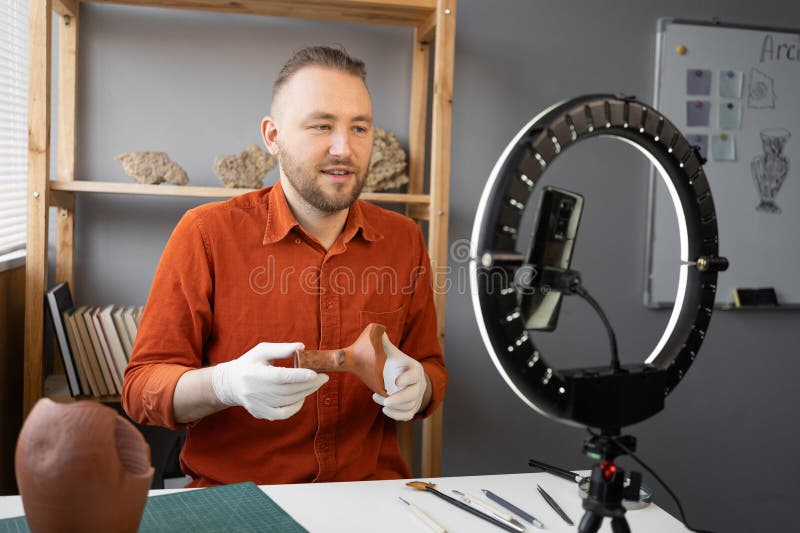 Archaeologist blogger broadcasts live with a ring lamp in his office working with ancient artifacts of prehistoric culture. Copy space. Archaeologist blogger broadcasts live with a ring lamp in his office working with ancient artifacts of prehistoric culture. Copy space