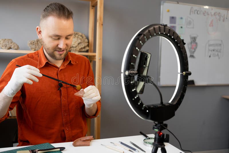 Archaeologist blogger broadcasts live with a ring lamp in his office, working with ancient artifacts of prehistoric culture, cleaning sand with a brush. Copy space. Archaeologist blogger broadcasts live with a ring lamp in his office, working with ancient artifacts of prehistoric culture, cleaning sand with a brush. Copy space