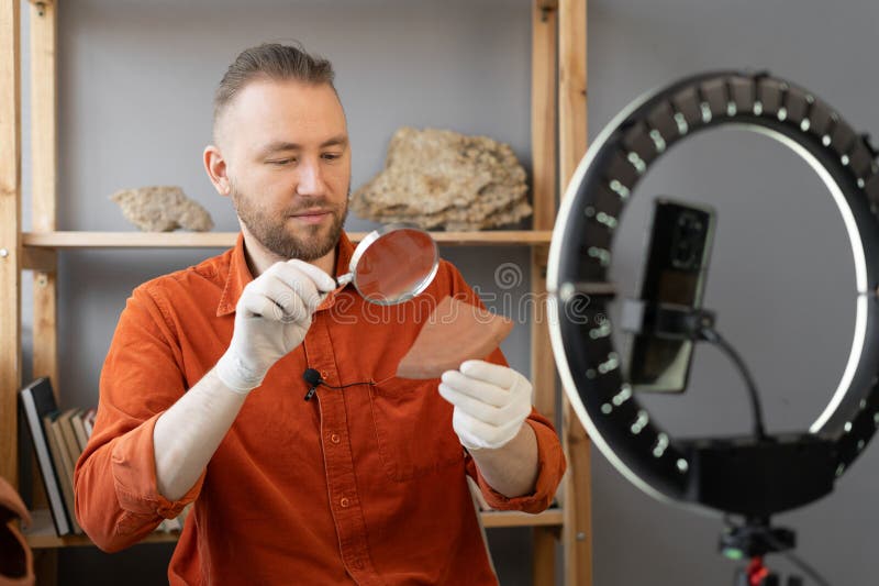 Archaeologist blogger broadcasts live with a ring lamp in his office working with ancient artifacts of prehistoric culture looks through a magnifying glass. Copy space. Archaeologist blogger broadcasts live with a ring lamp in his office working with ancient artifacts of prehistoric culture looks through a magnifying glass. Copy space