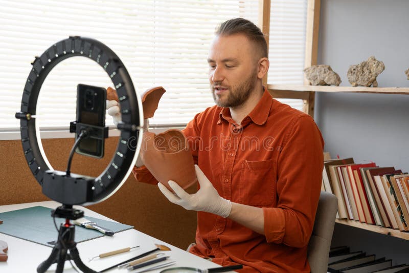 Archaeologist blogger broadcasts live with a ring lamp in his office working with ancient artifacts of prehistoric culture. Copy space. Archaeologist blogger broadcasts live with a ring lamp in his office working with ancient artifacts of prehistoric culture. Copy space