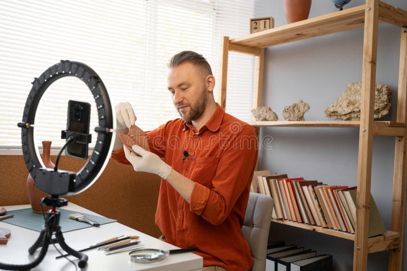 Archaeologist blogger broadcasts live with a ring lamp in his office working with ancient artifacts of prehistoric culture. Copy space. Archaeologist blogger broadcasts live with a ring lamp in his office working with ancient artifacts of prehistoric culture. Copy space