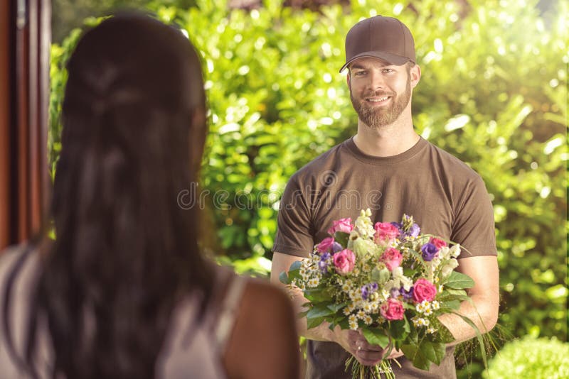 Smiling bearded 20s men wearing brown cap and brown t-shirt delivers flowers to door of young brunette female. Over the shoulder rear view of brunette. Smiling bearded 20s men wearing brown cap and brown t-shirt delivers flowers to door of young brunette female. Over the shoulder rear view of brunette.