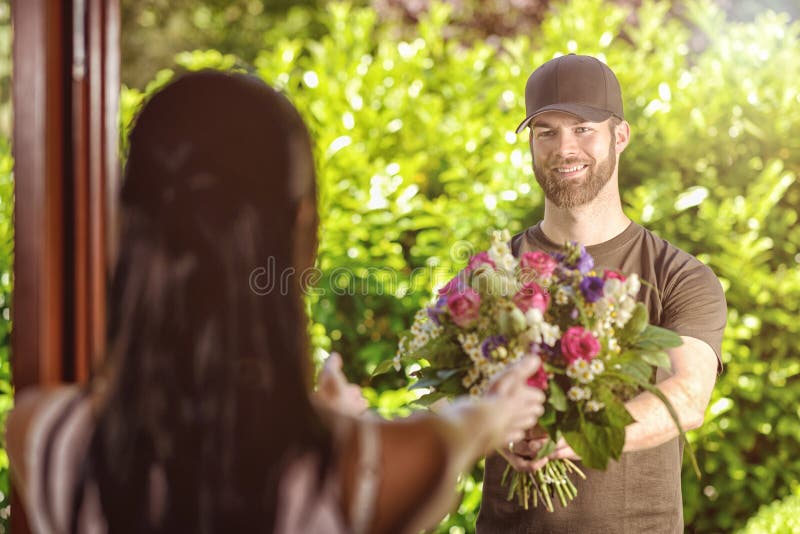 Smiling bearded 20s men wearing brown cap and brown t-shirt delivers flowers to door of young brunette female. Over the shoulder rear view of brunette. Smiling bearded 20s men wearing brown cap and brown t-shirt delivers flowers to door of young brunette female. Over the shoulder rear view of brunette.