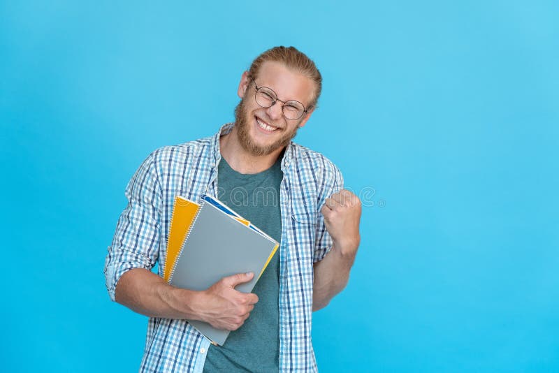 Bearded happy excited rejoicing man student hipster glasses hold copybooks notebooks yeah victory win gesture hand in fist smile success test exam I did it isolated blue studio background copy space. Bearded happy excited rejoicing man student hipster glasses hold copybooks notebooks yeah victory win gesture hand in fist smile success test exam I did it isolated blue studio background copy space.