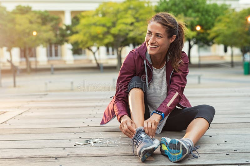 Mature fitness woman tie shoelaces on road. Cheerful runner sitting on floor on city streets with mobile and earphones wearing sport shoes. Active latin woman tying shoe lace before running. Mature fitness woman tie shoelaces on road. Cheerful runner sitting on floor on city streets with mobile and earphones wearing sport shoes. Active latin woman tying shoe lace before running.