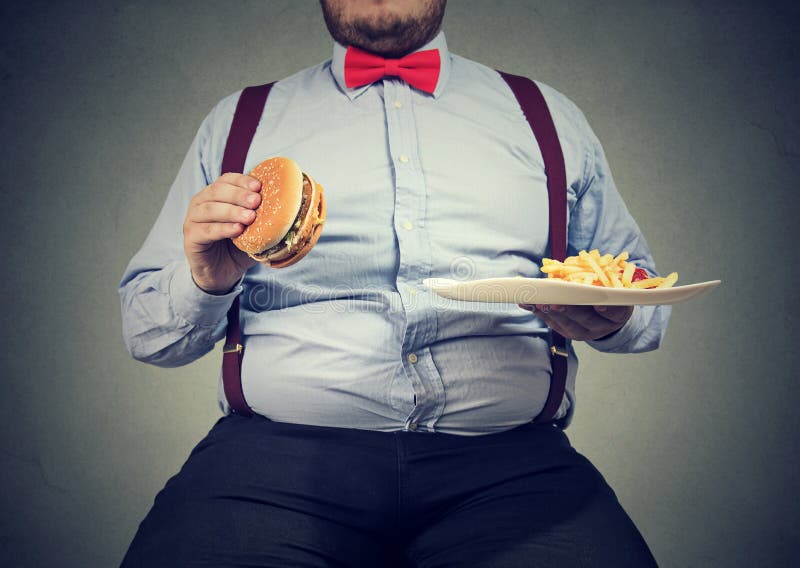 Crop shot of big man in formal clothes sitting and consuming plate with fast food on gray. Crop shot of big man in formal clothes sitting and consuming plate with fast food on gray.