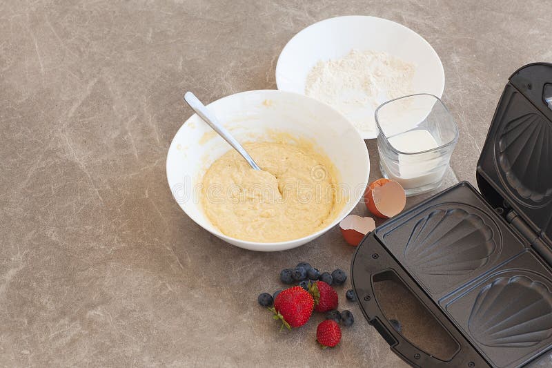 Cooking waffles at home - waffle iron, dough in a bowl, sugar and eggs on a marble background. Recipe step by step, step 2. Cooking waffles at home - waffle iron, dough in a bowl, sugar and eggs on a marble background. Recipe step by step, step 2