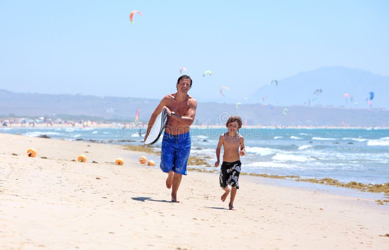 Happy young father and son running along busy, sandy kitesurfing beach with surfboard in the sun. Happy young father and son running along busy, sandy kitesurfing beach with surfboard in the sun