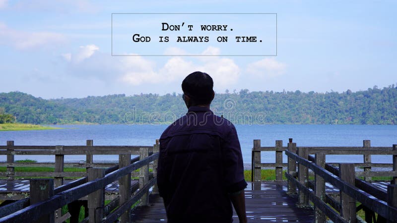 Inspirational quote - Don't worry. God is always on time. With a man standing alone from behind on wooden bridge looking at the lake view. Spiritual concept. Believe in God. Surrender to God. Inspirational quote - Don't worry. God is always on time. With a man standing alone from behind on wooden bridge looking at the lake view. Spiritual concept. Believe in God. Surrender to God