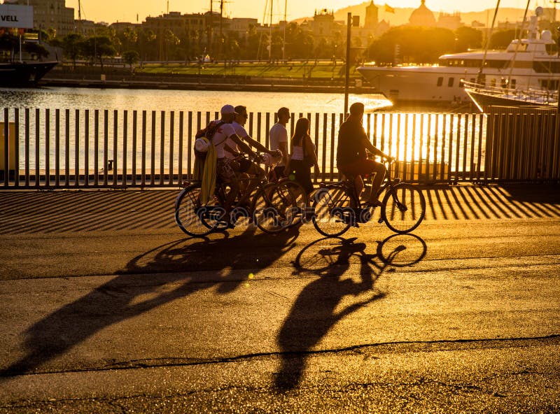 Group of cyclists beside the harbor at Port Vell, Barcelona, Spain with ships berthed and all seen in golden light at sunset. Group of cyclists beside the harbor at Port Vell, Barcelona, Spain with ships berthed and all seen in golden light at sunset.