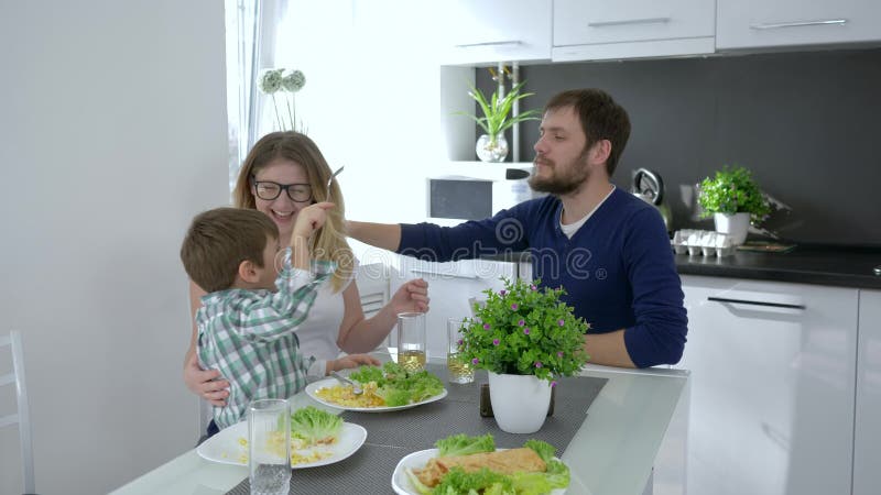 Вегетарианская семья, счастливые родители при малыш есть вкусную еду сидя на таблице в кухне