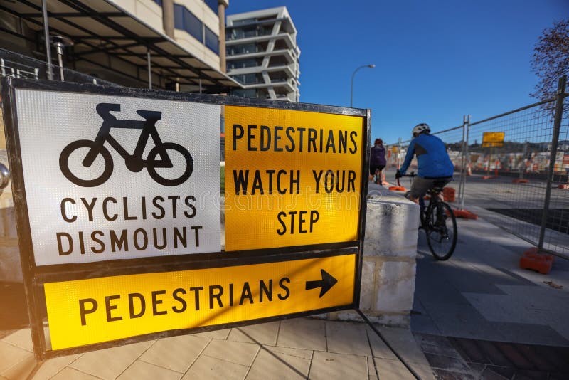 Cyclists dismount safety warning sign applying on public busy residential areas where construction worker repairing upgrading footpath with defocused Cyclists entering at the background, south Perth city CBD, Australia. Cyclists dismount safety warning sign applying on public busy residential areas where construction worker repairing upgrading footpath with defocused Cyclists entering at the background, south Perth city CBD, Australia