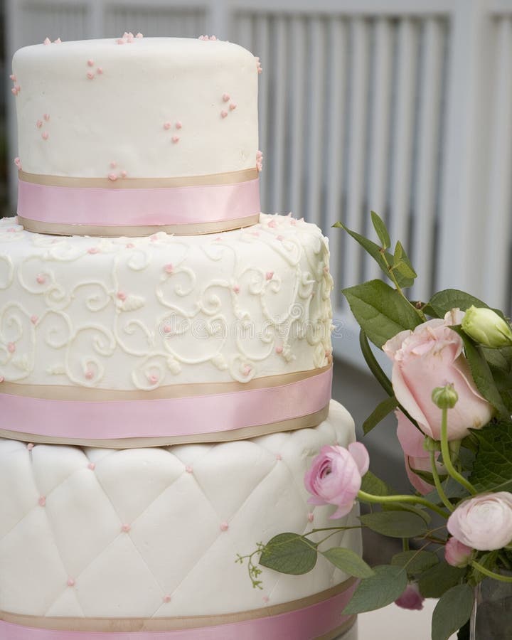 Close up of tiered white rolled fondant wedding cake, layers wrapped with pink and khaki ribbon decorated with quilted pattern, swirls and dots next to a pink fresh floral arrangement. Close up of tiered white rolled fondant wedding cake, layers wrapped with pink and khaki ribbon decorated with quilted pattern, swirls and dots next to a pink fresh floral arrangement