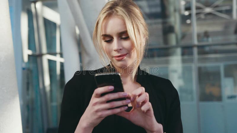 Взгляд вращения красивой молодой белокурой женщины в черной элегантной блузке используя ее smartphone в крупном аэропорте