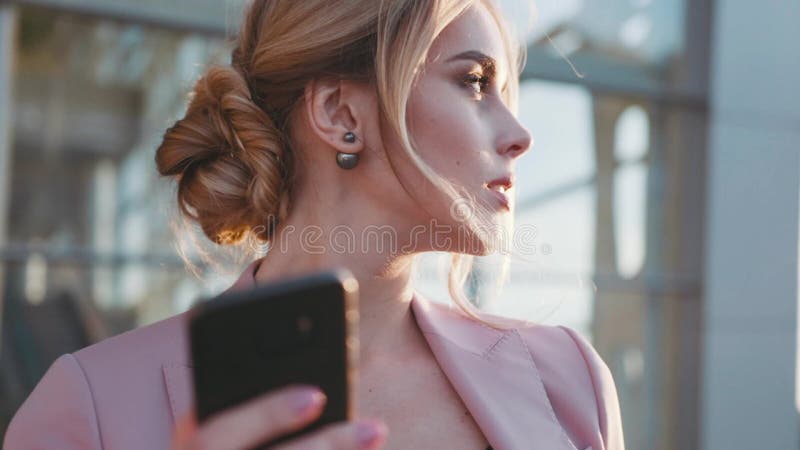 Взгляд вращения молодой белокурой дамы дела усмехаясь и используя ее телефон в ярком свете самомоднейшие технологии