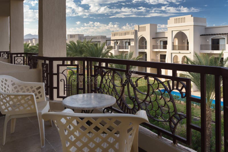 View at palm trees and a hotel foreside from a terrace with chairs and a table. Sunny day with blue sky and white clouds. Resort in Egypt. View at palm trees and a hotel foreside from a terrace with chairs and a table. Sunny day with blue sky and white clouds. Resort in Egypt.