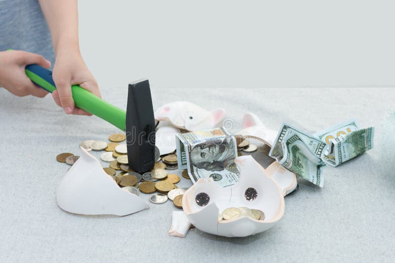 Close-up of children`s hands holding a hammer smash a piggy bank. Coins and hundred dollar bills spilled out of it. Horizontal photo, place for text. Close-up of children`s hands holding a hammer smash a piggy bank. Coins and hundred dollar bills spilled out of it. Horizontal photo, place for text