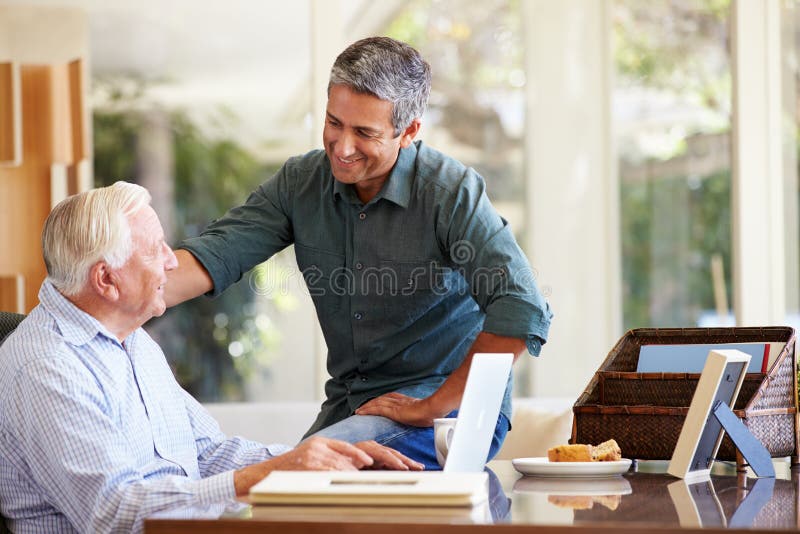 Adult Son Helping Father With Laptop At Home Putting Hand On Shoulder Smiling. Adult Son Helping Father With Laptop At Home Putting Hand On Shoulder Smiling