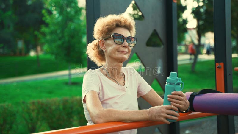 взрослая женщина отдыхает после занятий на уличной спортивной площадке, держа летом колбу с водой. пожилая женщина акции видеоматериалы