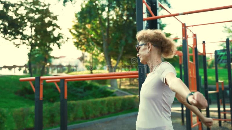 взрослая женщина разогревается перед тренировкой в спортзале летом. престарелые женщины проводят занятия по разогрев оружия и видеоматериал