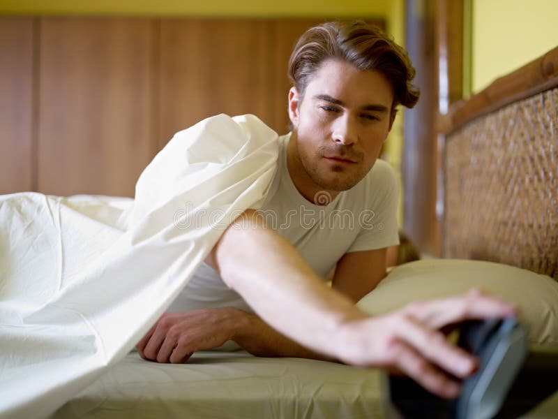 Caucasian adult man in bed snoozing alarm clock and waking up. Horizontal shape, waist up, front view. Caucasian adult man in bed snoozing alarm clock and waking up. Horizontal shape, waist up, front view