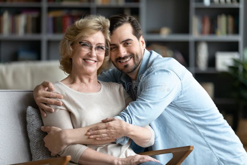 Younger and older generation relatives people portrait, love, care, connection, family bonds concept. Grown up adult son hugs elderly mother while she resting seated on cozy comfort armchair indoors. Younger and older generation relatives people portrait, love, care, connection, family bonds concept. Grown up adult son hugs elderly mother while she resting seated on cozy comfort armchair indoors