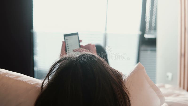 Вид сзади женщины брюнет лежа на кровати и просматривает интернет на smartphone Девушка тратя время дома видеоматериал Вид сзади женщины брюнет лежа на кровати и просматривает интернет на smartphone Девушка тратя время дома видеоматериал