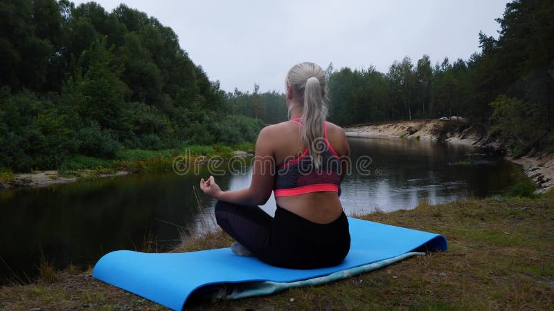 вид с задней стороны красивой блондинки, которая делает йогу на краю обрыва возле реки. молодая женщина сидит в