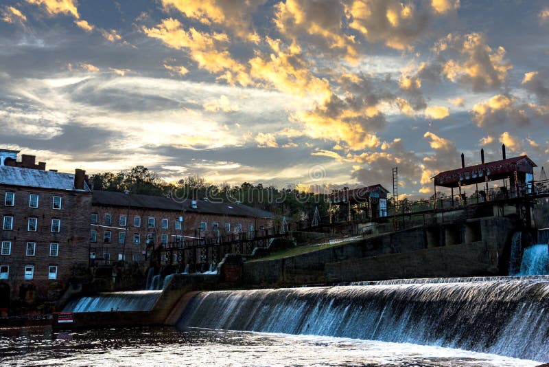 Prattville, Alabama, USA - December 20, 2016: Waterfall at the old mill pond with the Continental Gin Company in the background with a dramatic sunset. Prattville, Alabama, USA - December 20, 2016: Waterfall at the old mill pond with the Continental Gin Company in the background with a dramatic sunset.