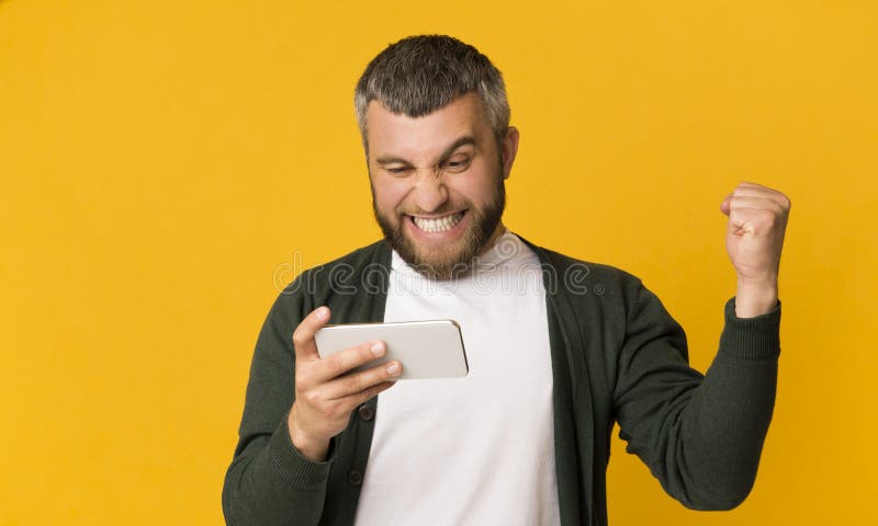 Возбужденный бородатый смартфон удерживания человека и обхватывая кулак стоковые фото