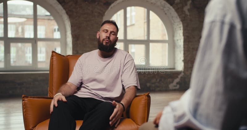возбужденный мужчина с бородой в розовой рубашке рассказывает о своих проблемах и вопросах психологу. сидит молодой человек