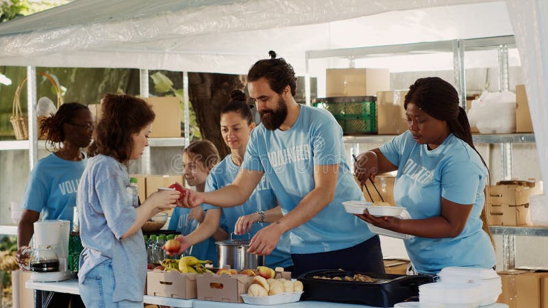 Young people united by commitment to charity, aiding underprivileged and contributing food to individuals experiencing homelessness. Volunteers sharing free meals to the poor and needy. Tripod shot. Young people united by commitment to charity, aiding underprivileged and contributing food to individuals experiencing homelessness. Volunteers sharing free meals to the poor and needy. Tripod shot.