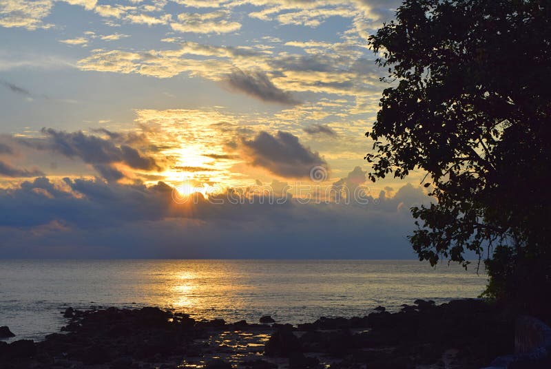 This is a photograph of rising sun with golden sunshine at horizon.. The clouds are lined with golden lines and there are silhouettes of tree and stones... The image is captured at Laxmanpur, Neil Island, Andaman Nicobar islands, India. This is a photograph of rising sun with golden sunshine at horizon.. The clouds are lined with golden lines and there are silhouettes of tree and stones... The image is captured at Laxmanpur, Neil Island, Andaman Nicobar islands, India...